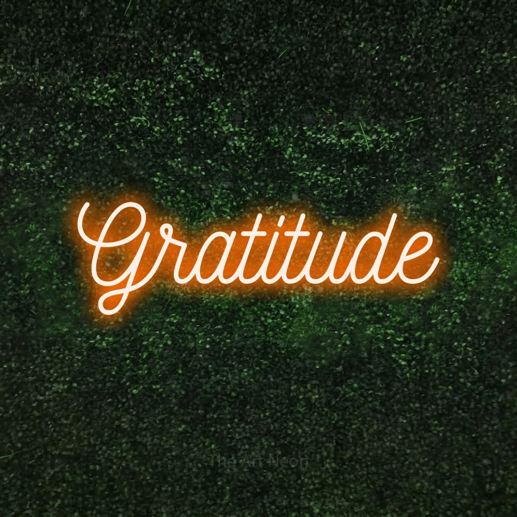 Orange script-style LED neon sign reading "Gratitude" against a textured green moss wall