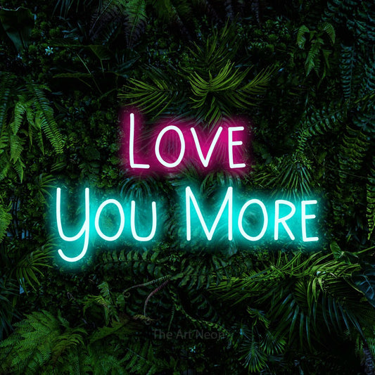 Ice blue and hot pink LED neon sign reading "Love You More" against a dense tropical foliage wall