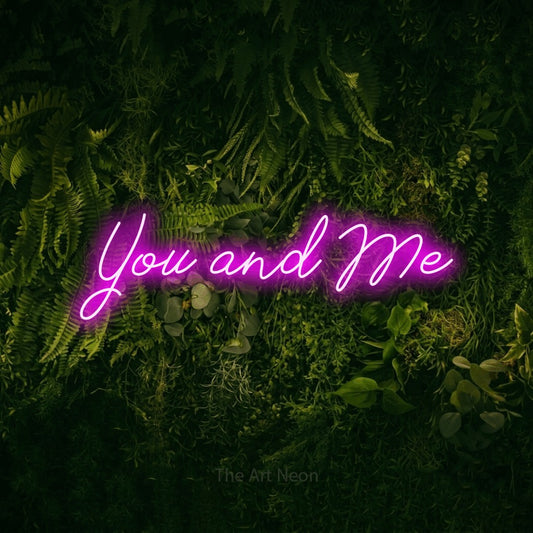 Pink script-style LED neon sign reading "You and Me" against a moss-covered green wall