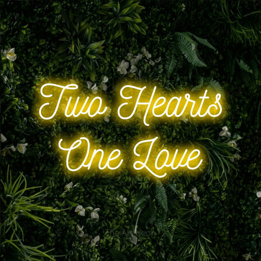 Yellow script-style LED neon sign reading "Two Hearts One Love" against a leafy green wall