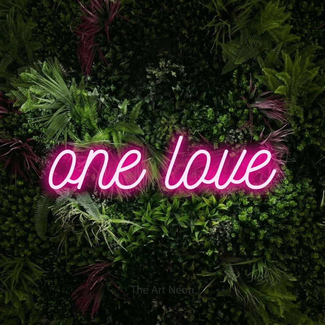 Enseigne néon à DEL à lumière rose vif, de style manuscrit, affichant « One Love » sur un mur de jardin luxuriant aux fleurs roses