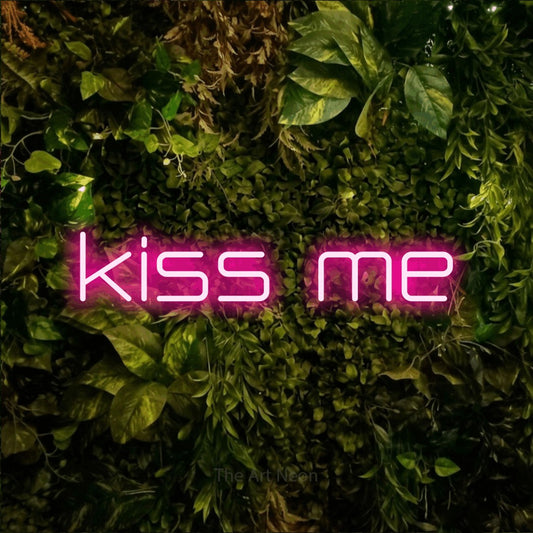 Hot pink outline-style LED neon sign reading "Kiss Me" against a textured moss and foliage wall