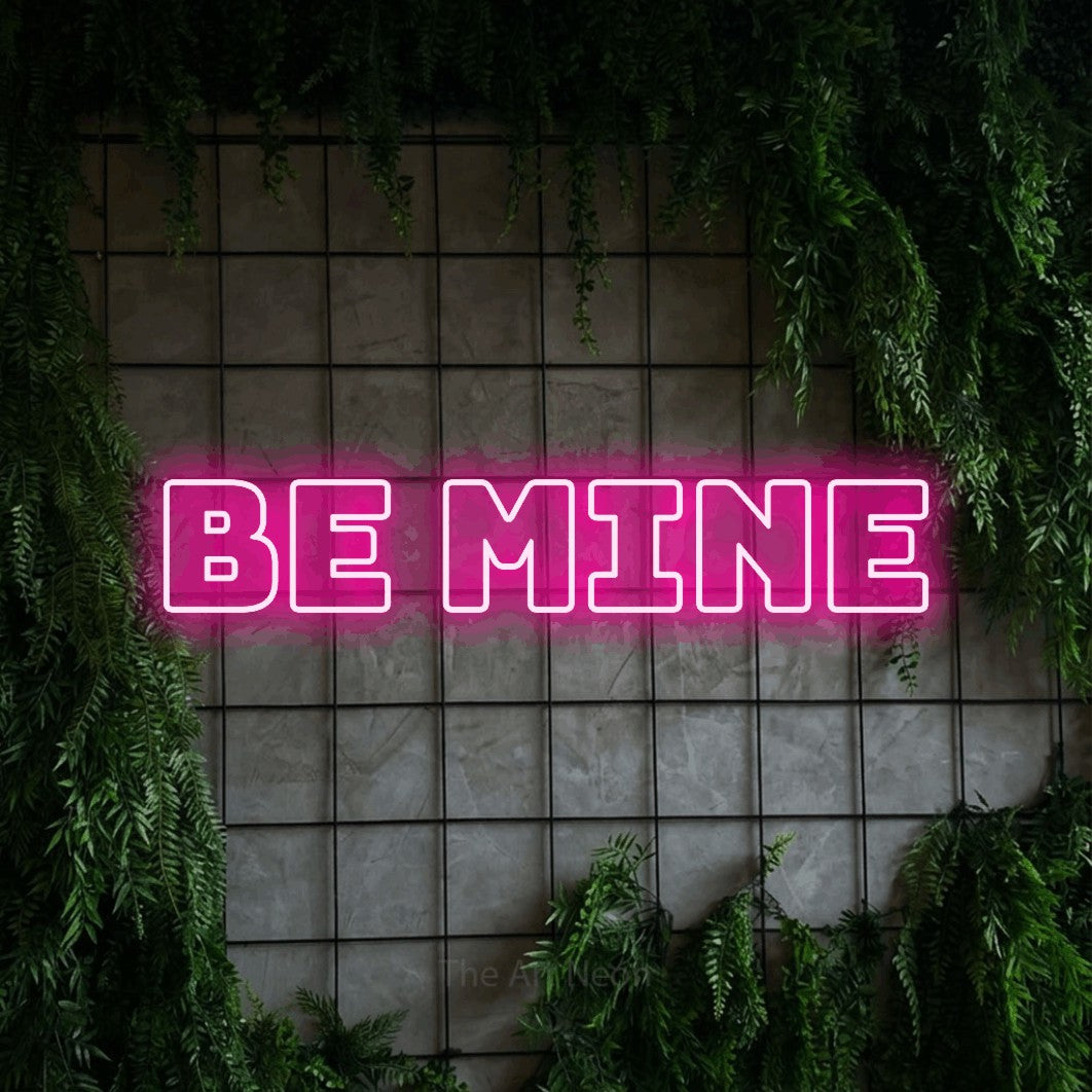 Hot pink outline-style LED neon sign reading "Be Mine" against a green grid wall with foliage