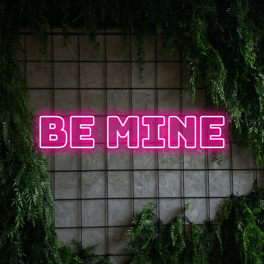 Hot pink outline-style LED neon sign reading "Be Mine" against a green grid wall with foliage