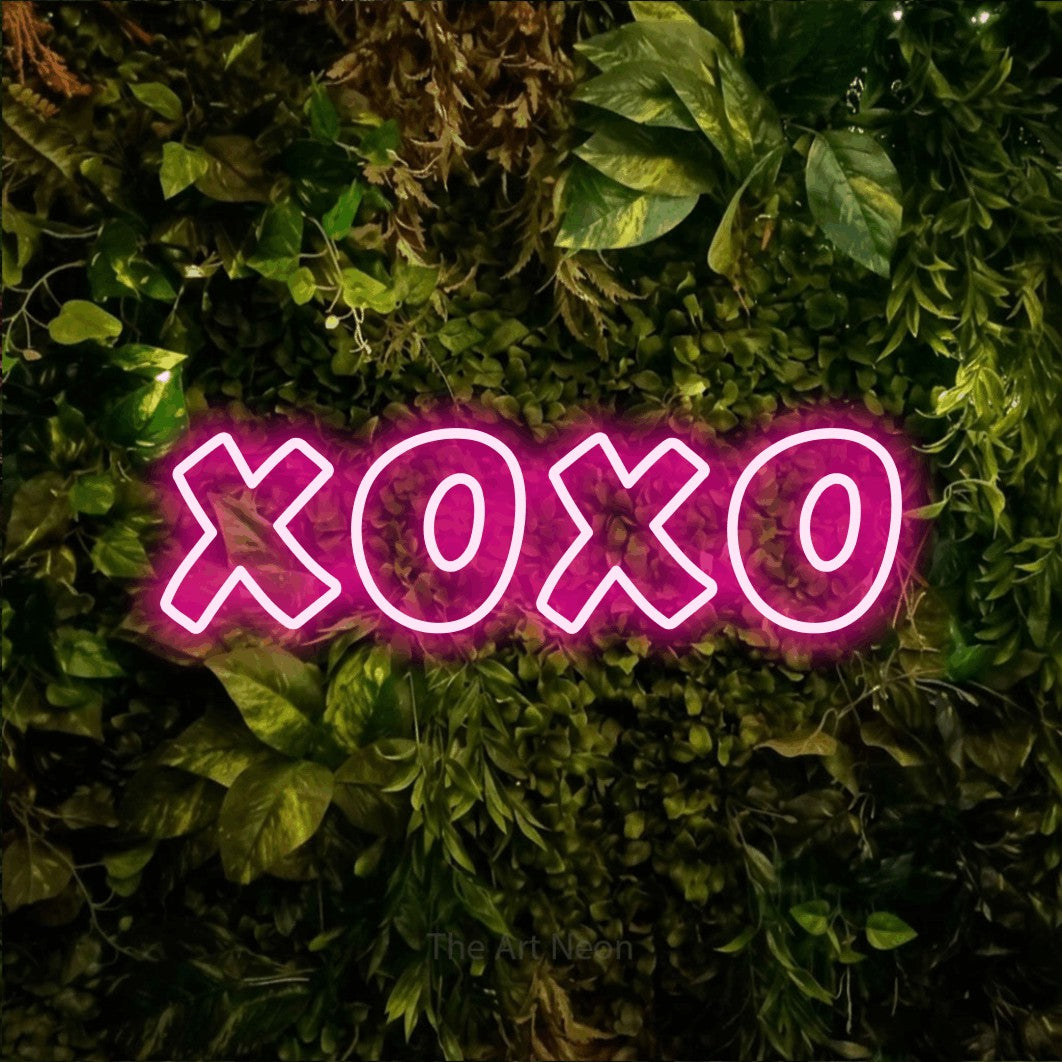 Hot pink outline-style LED neon sign reading "XOXO" against a textured moss and foliage wall