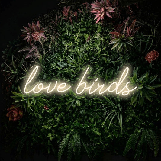 Warm white script-style LED neon sign reading "Love Birds" against a flowering garden wall
