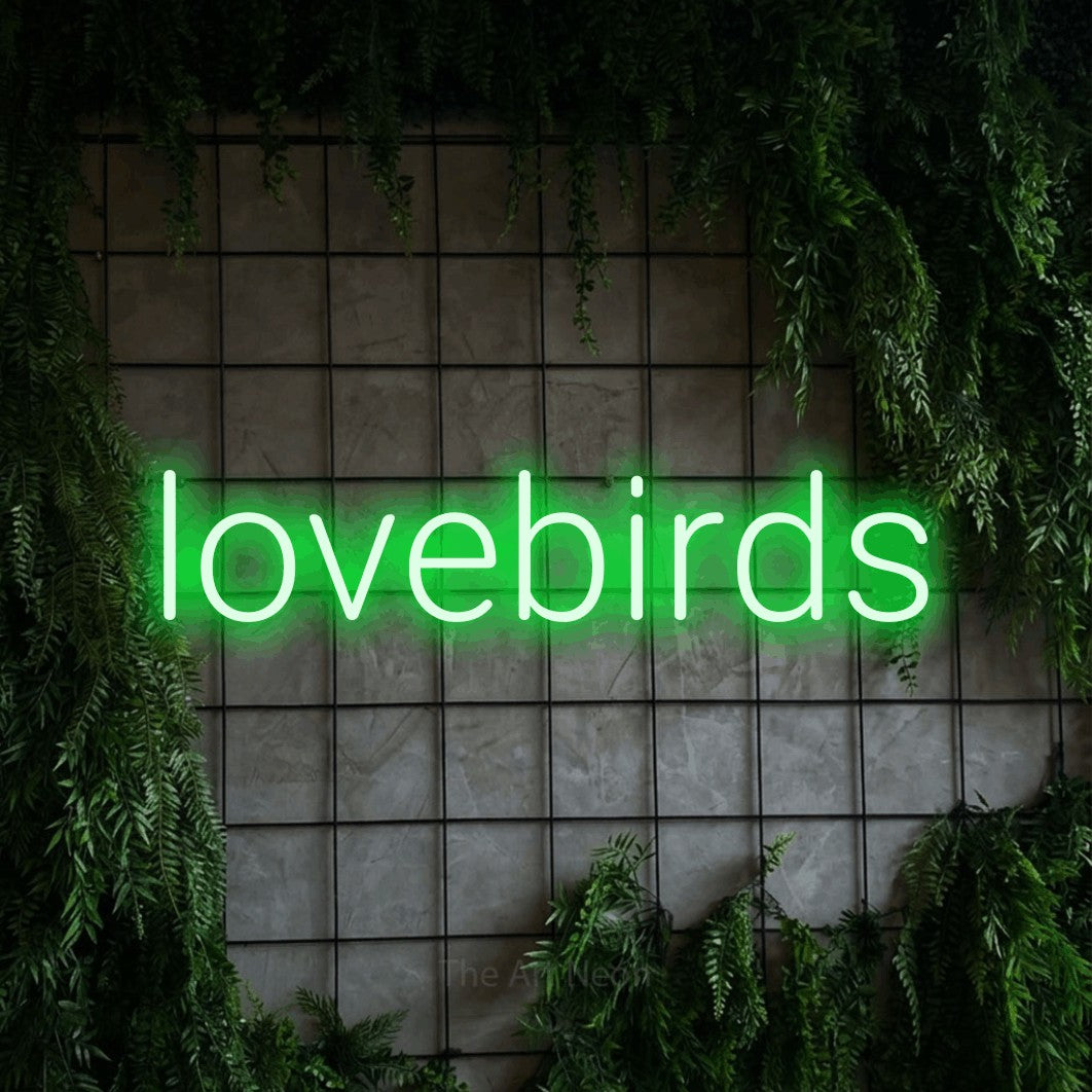 Green LED neon sign reading "Lovebirds" against a green grid wall with foliage