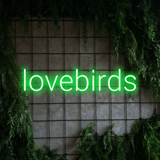 Green LED neon sign reading "Lovebirds" against a green grid wall with foliage
