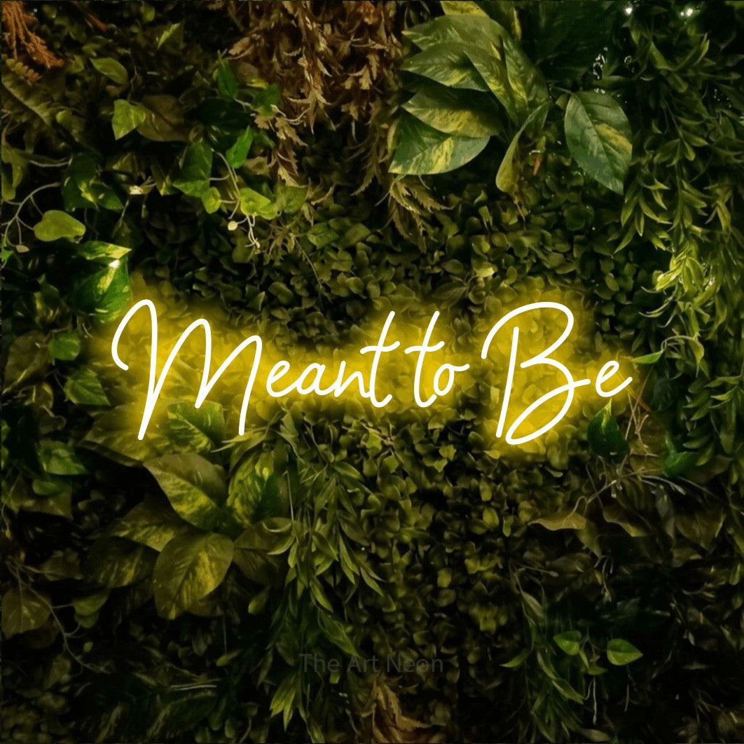 Yellow script-style LED neon sign reading "Meant to Be" against a textured moss and foliage wall