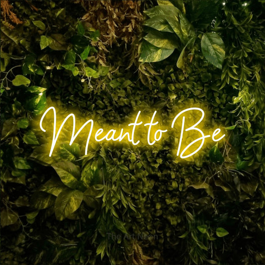 Yellow script-style LED neon sign reading "Meant to Be" against a textured moss and foliage wall