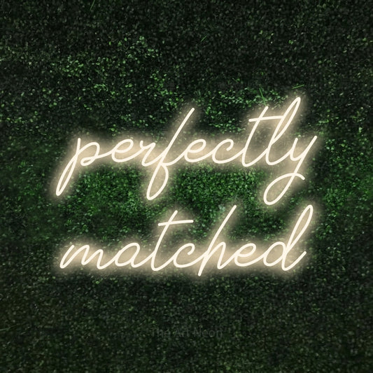 Warm white script-style LED neon sign reading "Perfectly Matched" against a textured green moss wall