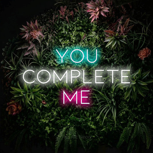 Multicolour LED neon sign reading "You Complete Me" against a flowering garden wall