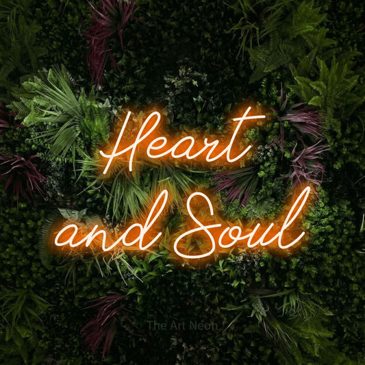 Orange script-style LED neon sign reading "Heart and Soul" against a lush garden wall with pink flowers