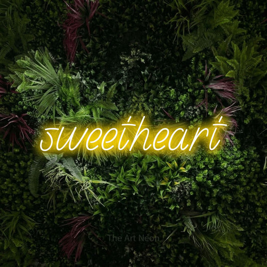 Yellow LED neon sign reading "Sweetheart" against a lush garden wall with pink flowers
