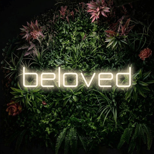 Warm white LED neon sign reading "Beloved" against a flowering garden wall