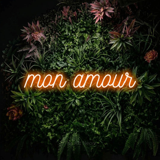 Orange script-style LED neon sign reading "Mon Amour" against a flowering garden wall