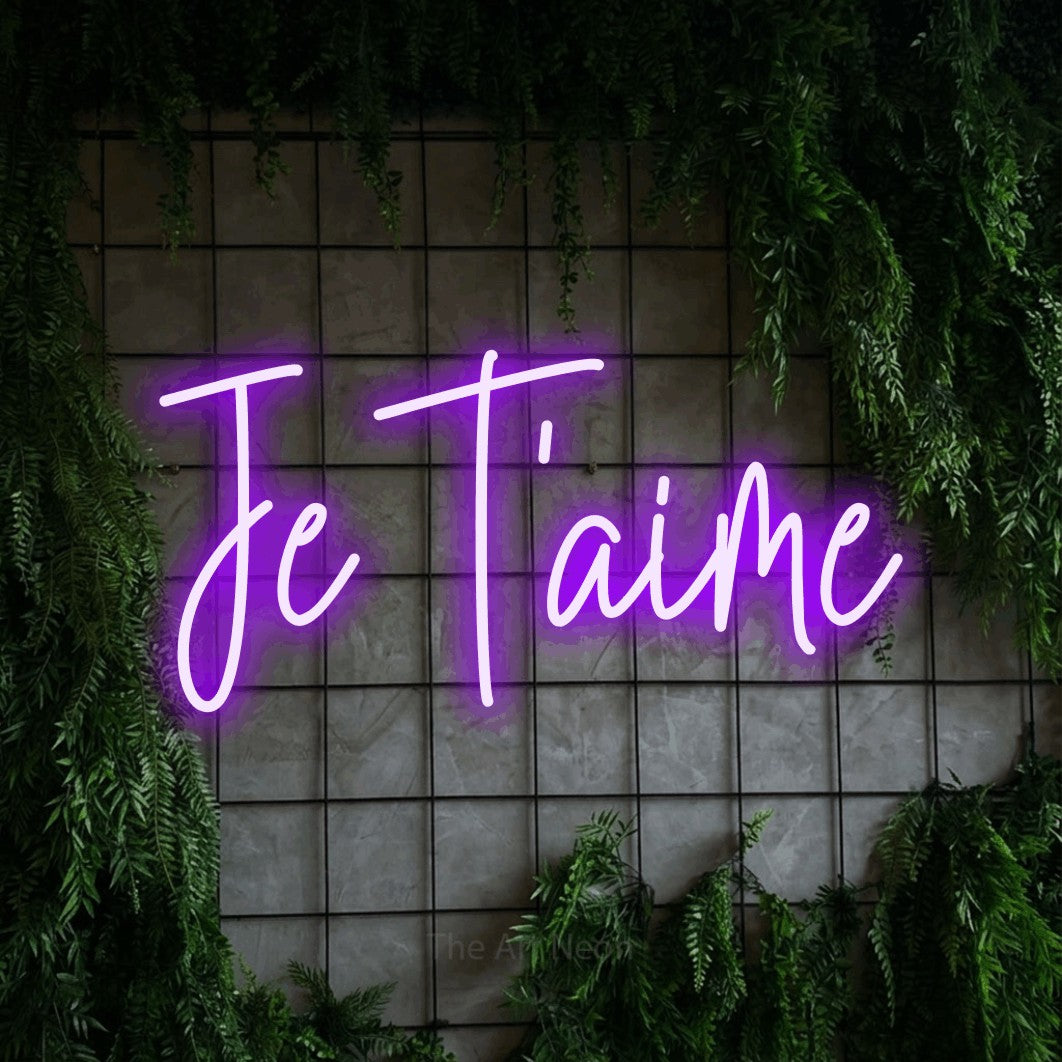 Purple script-style LED neon sign reading "Je T'aime" against a green grid wall with foliage