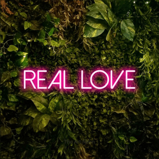 Hot pink LED neon sign reading "Real Love" against a textured moss and foliage wall
