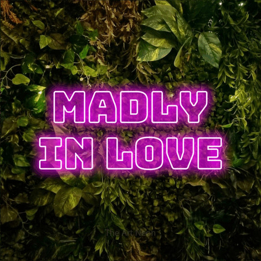 Pink outline-style LED neon sign reading "Madly in Love" against a textured moss and foliage wall