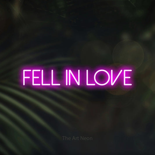 Pink LED neon sign reading "Fell in Love" against dark blurred palm leaves