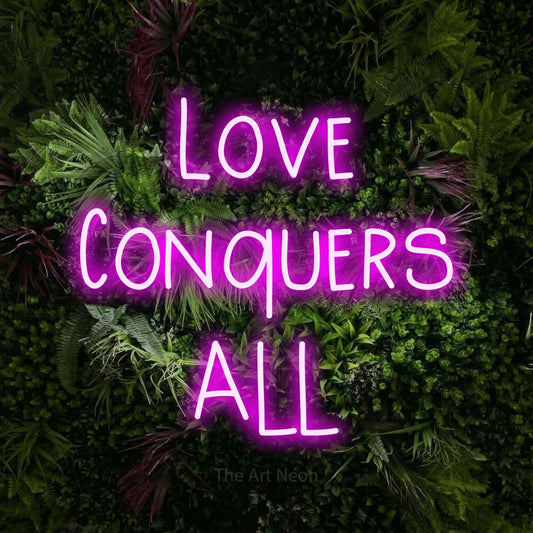 Pink LED neon sign reading "Love Conquers All" against a lush garden wall with pink flowers