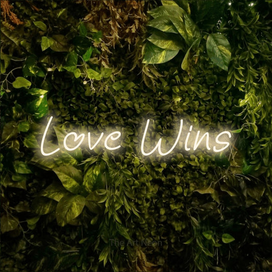Warm white LED neon sign reading "Love Wins" against a textured moss and foliage wall