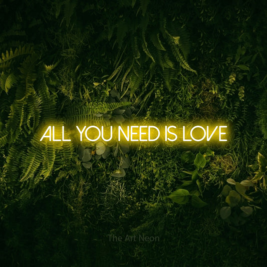 Yellow LED neon sign reading "All You Need is Love" against a moss-covered green wall