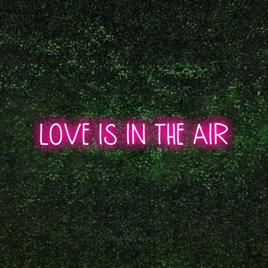 Hot pink LED neon sign reading "Love is in the Air" against a textured green moss wall