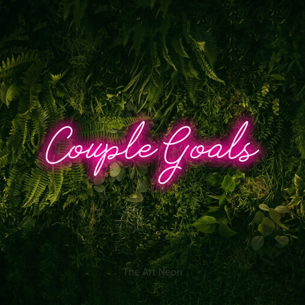 Hot pink script-style LED neon sign reading "Couple Goals" against a moss-covered green wall