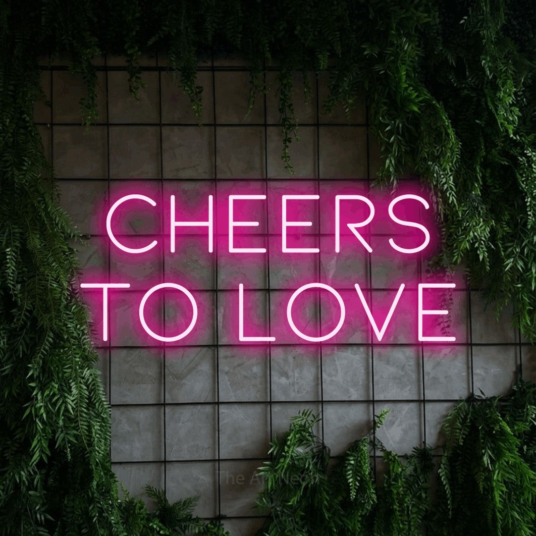 Hot pink LED neon sign reading "Cheers to Love" against a green grid wall with foliage
