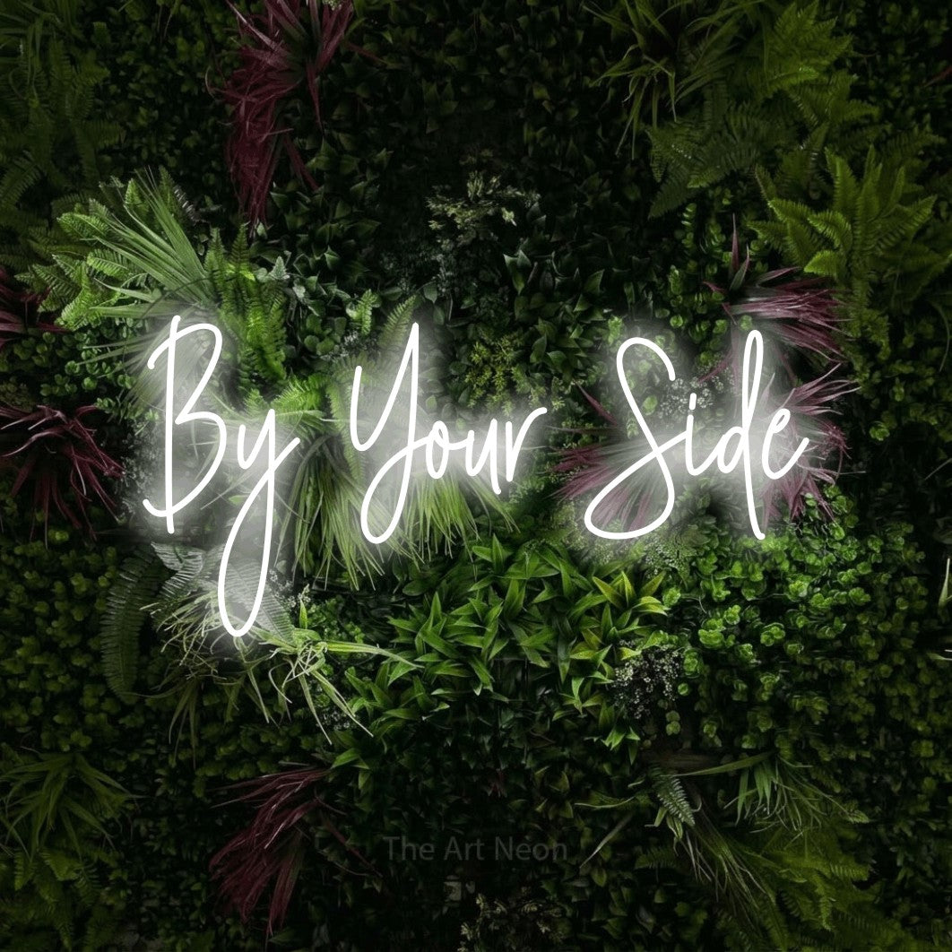 Cool white script-style LED neon sign reading "By Your Side" against a lush garden wall with pink flowers