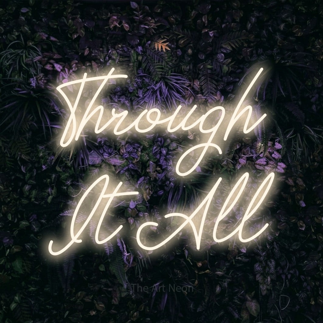 Warm white script-style LED neon sign reading "Through It All" against a purple floral wall