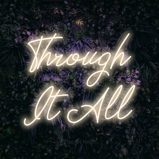 Warm white script-style LED neon sign reading "Through It All" against a purple floral wall