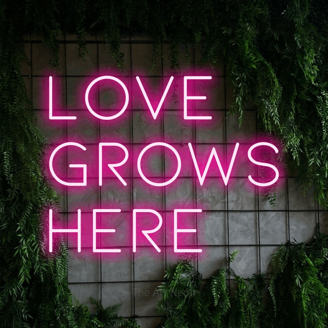 Hot pink LED neon sign reading "Love Grows Here" against a green grid wall with foliage