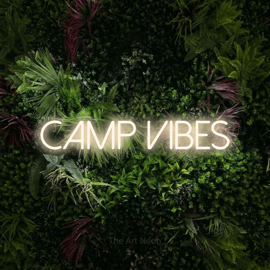 Warm white LED neon sign reading "Camp Vibes" against a lush garden wall with pink flowers