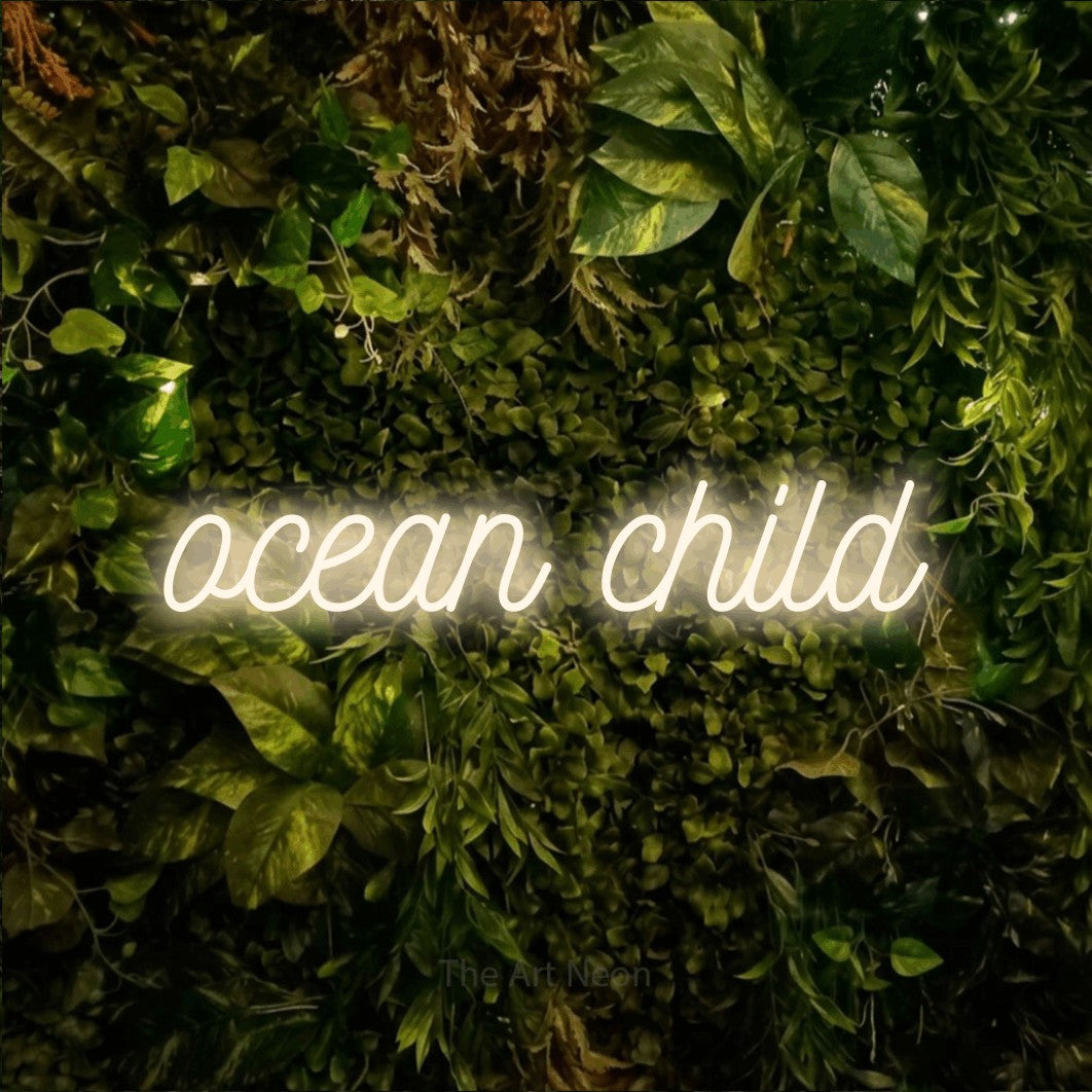 Warm white script-style LED neon sign reading "Ocean Child" against a textured moss and foliage wall