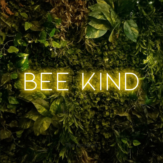 Yellow LED neon sign reading "Bee Kind" against a textured moss and foliage wall