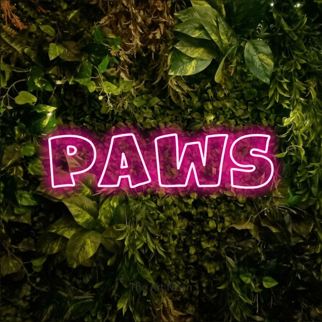 Hot pink outline-style LED neon sign reading "Paws" against a textured moss and foliage wall