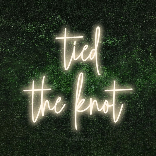 Warm white script-style LED neon sign reading "Tied the Knot" against a textured green moss wall for wedding décor