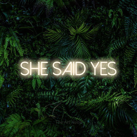 Warm white LED neon sign reading "She Said Yes" against a dense tropical foliage wall for wedding décor