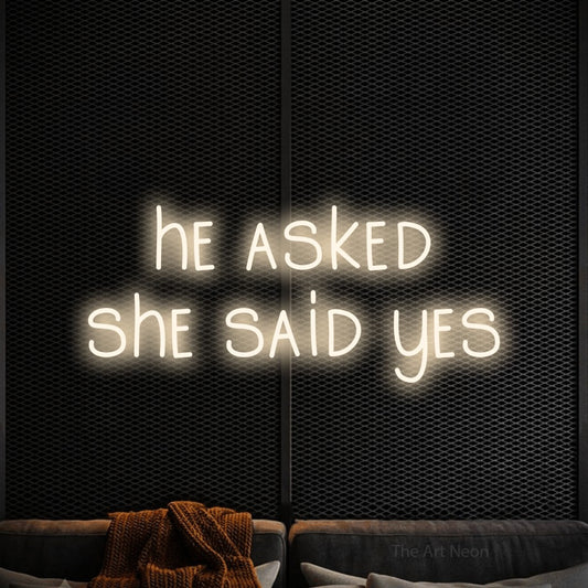 Warm white LED neon sign reading "He Asked She Said Yes" against a black textured wall for wedding décor