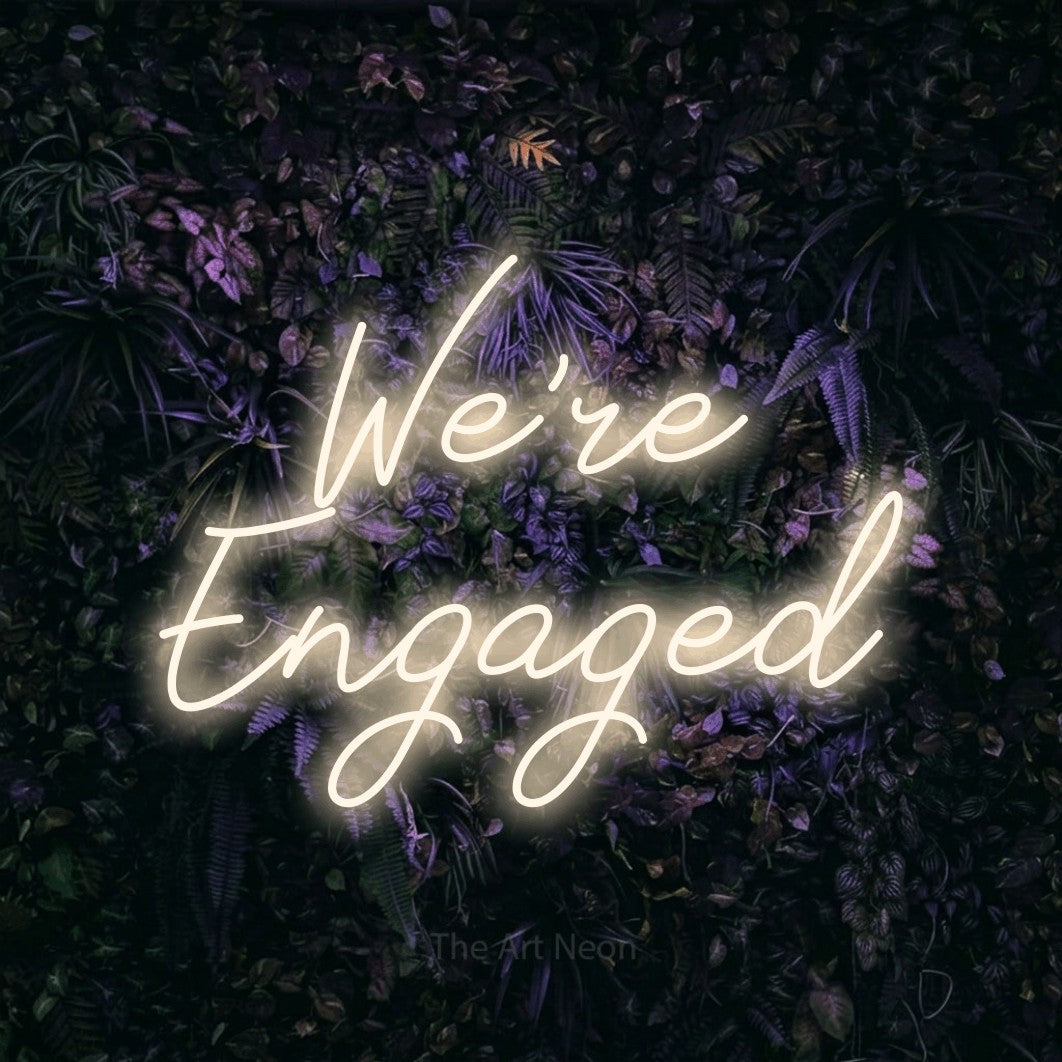 Warm white script-style LED neon sign reading "We're Engaged" against a purple floral wall for wedding décor