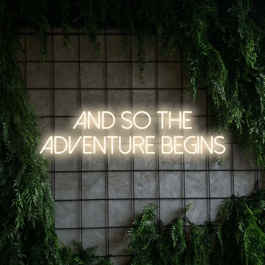 Warm white LED neon sign reading "And So the Adventure Begins" against a green grid wall with foliage for wedding décor