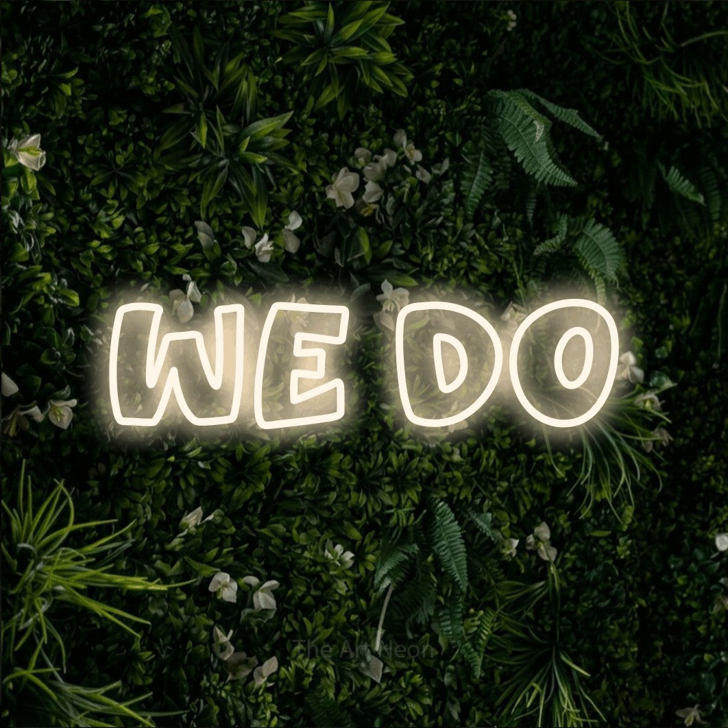 Warm white outline-style LED neon sign reading "We Do" against a leafy green wall for wedding décor