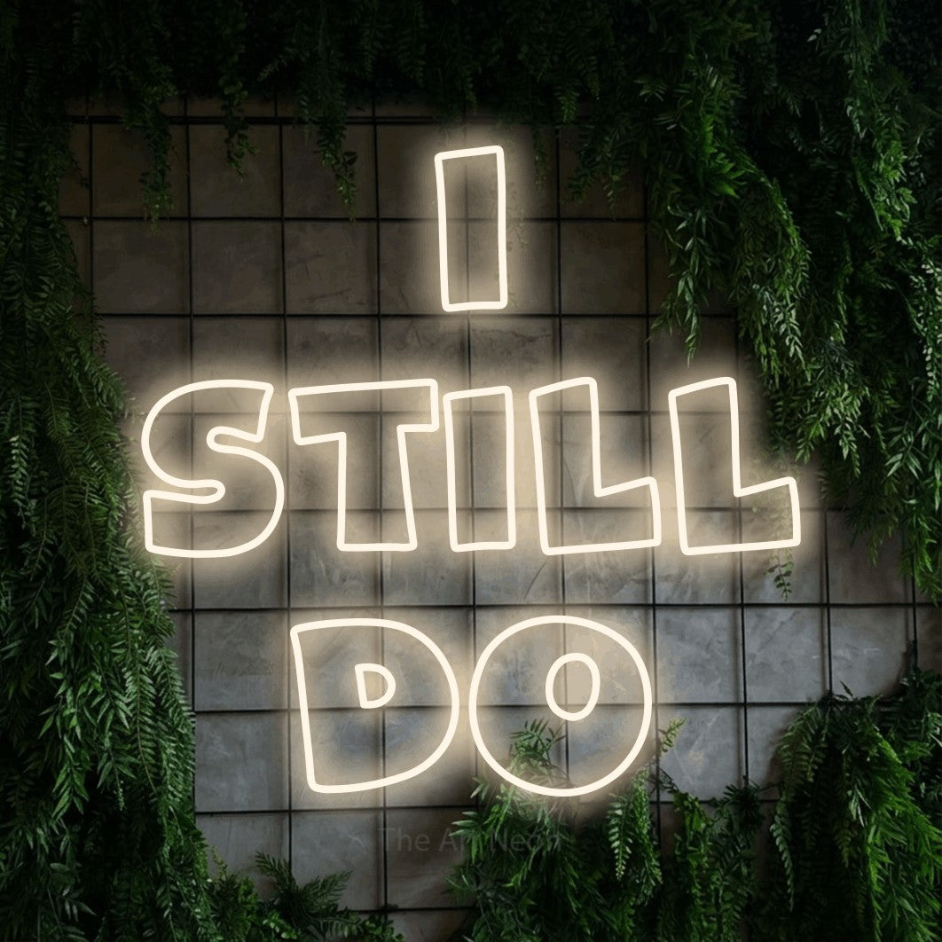 Warm white outline-style LED neon sign reading "I Still Do" against a green grid wall with foliage for wedding décor