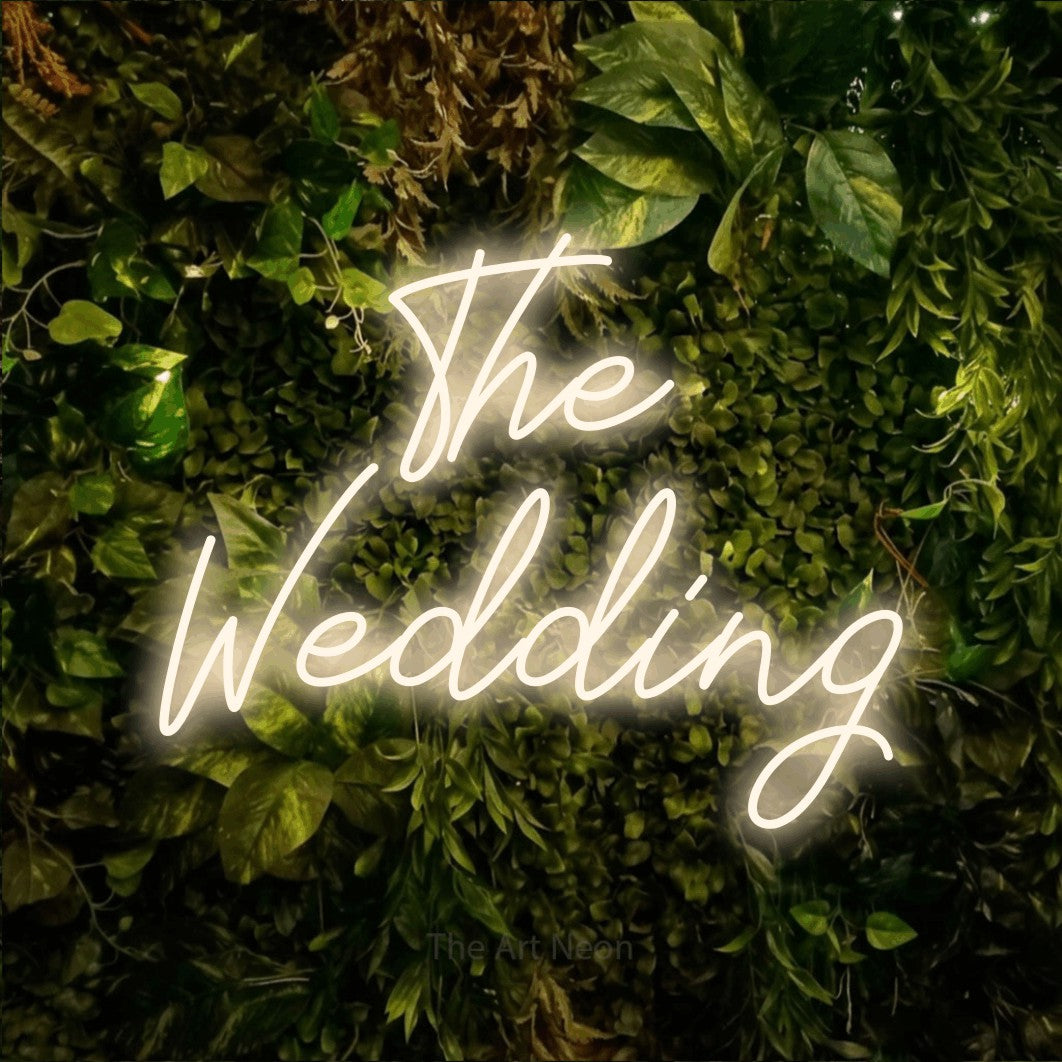 Warm white script-style LED neon sign reading "The Wedding" against a textured moss and foliage wall for wedding décor