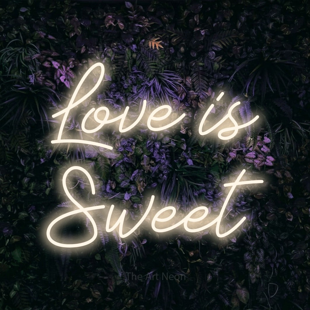 Warm white script-style LED neon sign reading "Love is Sweet" against a purple floral wall for wedding décor