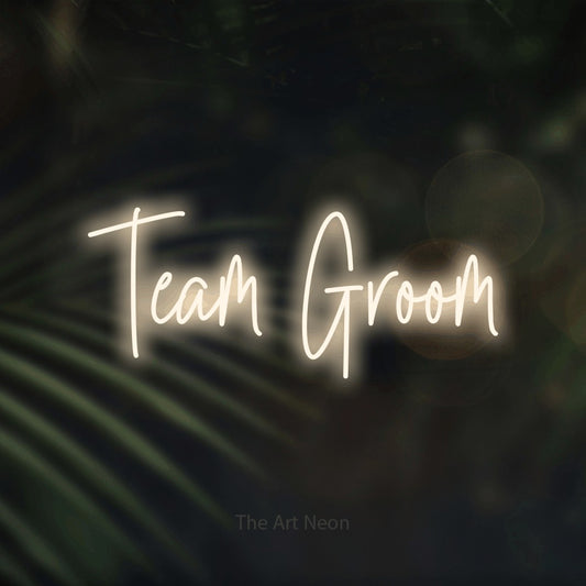 Warm white script-style LED neon sign reading "Team Groom" against dark blurred palm leaves for wedding décor