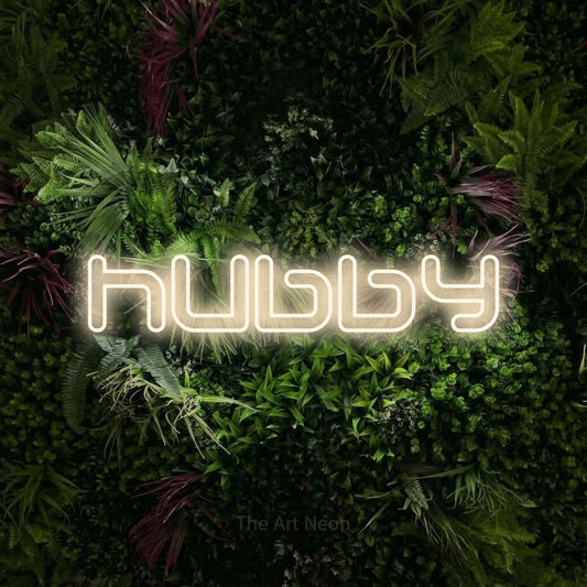 Warm white outline-style LED neon sign reading "Hubby" against a lush garden wall with pink flowers for wedding décor