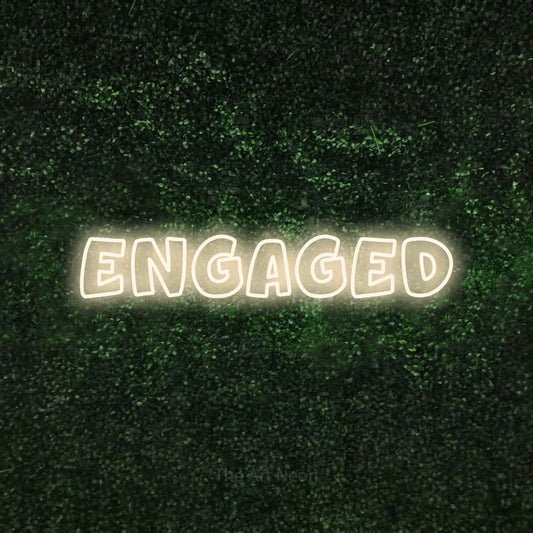 Warm white outline-style LED neon sign reading "Engaged" against a textured green moss wall for wedding décor